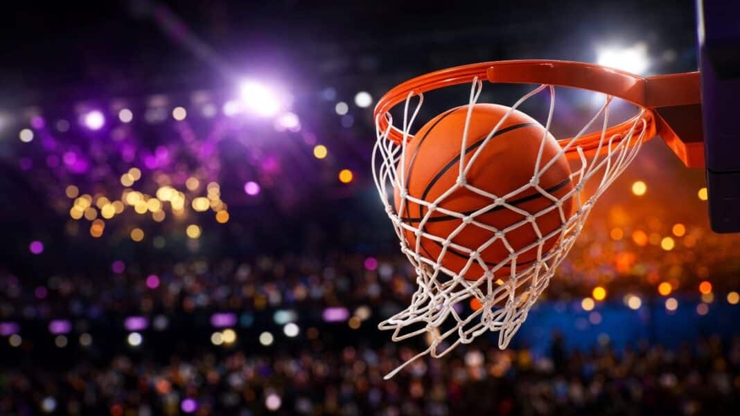 nba-sezona-zacetek-2026 Košarkarska žoga leti skozi obroč v polni dvorani, osvetljeni v vijolično-zlatih in oranžno-modrih barvah